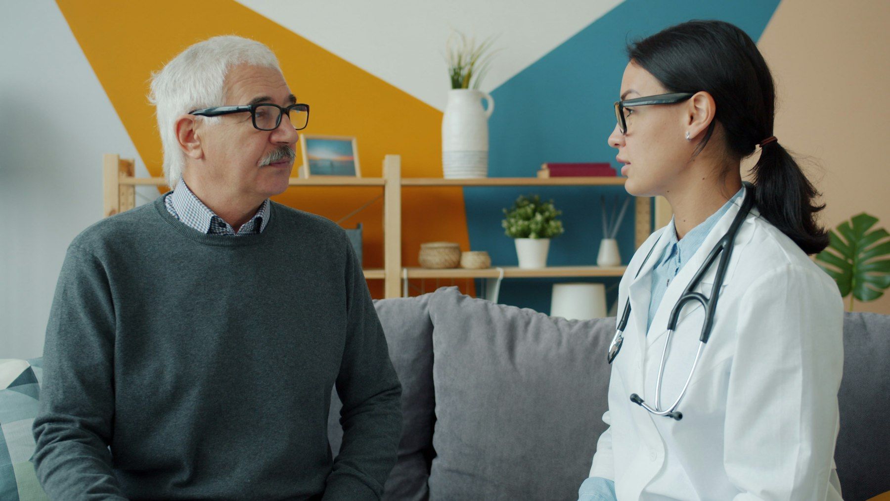 A doctor in a white coat talks with an older man on his couch